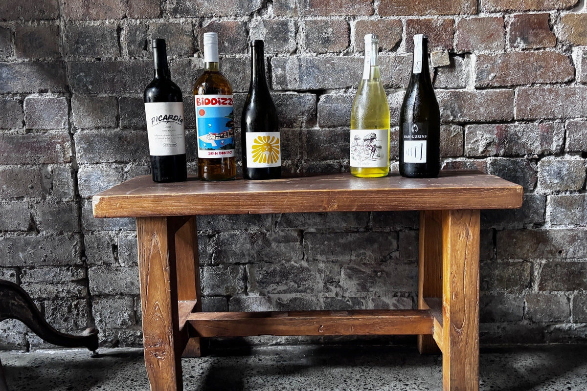 Pet nat and skin contact bottles from the Coogee Wine Room list, including Biddizza, See Saw 'The Party' and San Lurins, on a timber bench against a brick wall